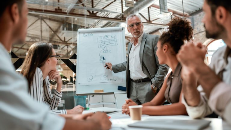 An experienced professional leads a diverse team in a collaborative discussion, explaining concepts on a flipchart, emphasizing the theme of 'Leadership Across Cultures'